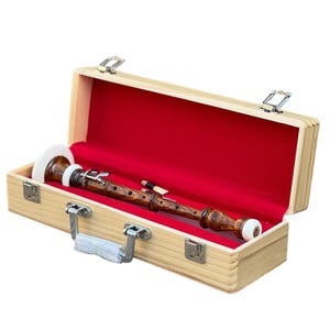 May include: A wooden recorder in a light-colored wooden case with a red velvet interior. The recorder is dark brown with white and light-colored accents. The case has metal latches and a handle.