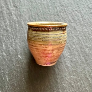 May include: A small, brown and tan ceramic cup with a striped pattern. The cup is sitting on a gray surface.