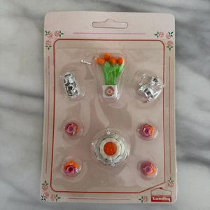 May include: A package of miniature dollhouse accessories, including a camera, a bouquet of orange tulips, a silver figurine, a cake, and several pink and orange roses. The items are arranged on a cardboard backing with a floral border. The brand name "Lundby" is visible.