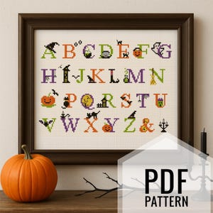 May include: A framed cross-stitch sampler featuring the alphabet with Halloween-themed designs. The letters are in various colors, with images of pumpkins, bats, and witches' hats. A bright orange pumpkin sits in front of the frame. The text "PDF PATTERN" is visible.
