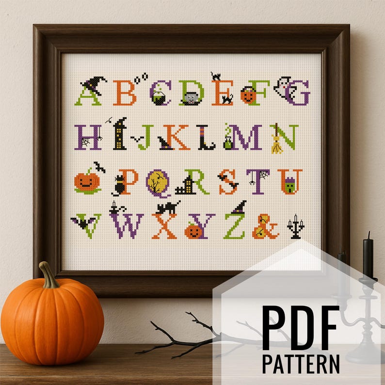 May include: A framed cross-stitch sampler featuring the alphabet with Halloween-themed designs. The letters are in various colors, with images of pumpkins, bats, and witches' hats. A bright orange pumpkin sits in front of the frame. The text "PDF PATTERN" is visible.