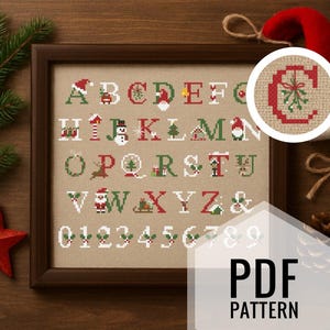 Puede incluir: Un muestrario de punto de cruz enmarcado con un alfabeto y números navideños, con diseños festivos como gorros de Papá Noel, muñecos de nieve y adornos. Una estrella roja, ramitas de pino y un gorro de Papá Noel están dispuestos alrededor del marco. El texto "PDF PATTERN" es visible.