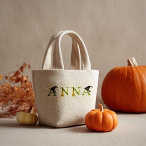May include: A beige canvas tote bag with the name "ANNA" embroidered in green and black, featuring witch hats and broomstick designs. Two pumpkins, one orange and one white, are next to the bag, suggesting a Halloween theme.