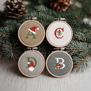 Peut inclure: Quatre petits ornements ronds en point de croix dans des cerceaux en bois. Chacun présente une lettre et un motif sur le thème de Noël : un "A" avec un bonnet de Père Noël, un "C" avec du gui, un "D" avec un gnome et un "B" avec un cadeau. Le fond comprend des aiguilles de pin.