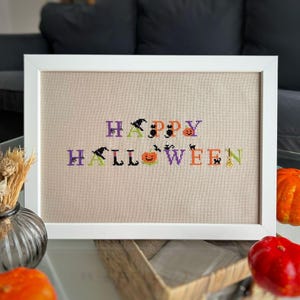 May include: A framed cross-stitch artwork with the words "HAPPY HALLOWEEN" in purple, orange, black, and green. The letters are decorated with witch hats, bats, and pumpkins. The artwork is in a white frame.