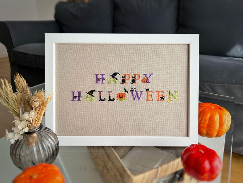 May include: A framed cross-stitch artwork with the words "HAPPY HALLOWEEN" in purple, orange, black, and green. The letters are decorated with witch hats, bats, and pumpkins. The artwork is in a white frame.