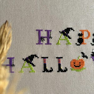 May include: Cross-stitch design on beige fabric spelling "HAPPY HALLOWEEN". The letters are in purple, green, orange, and black. The design includes a witch hat, a black cat, a pumpkin, and a bat.