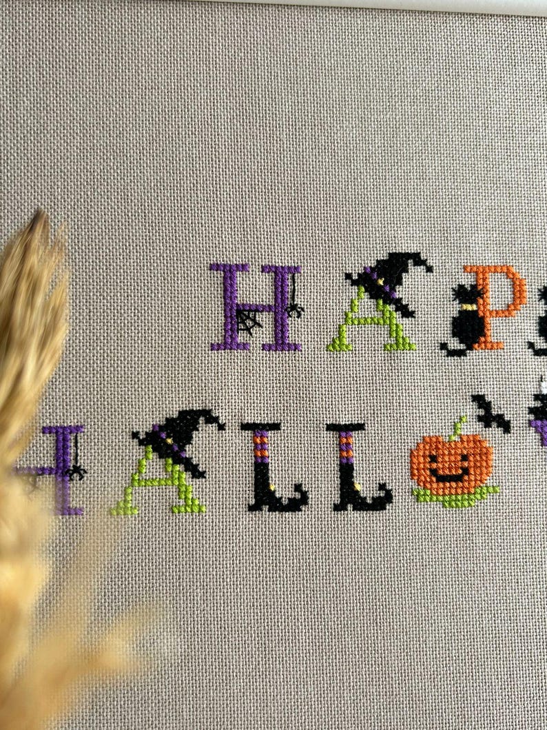 May include: Cross-stitch design on beige fabric spelling "HAPPY HALLOWEEN". The letters are in purple, green, orange, and black. The design includes a witch hat, a black cat, a pumpkin, and a bat.
