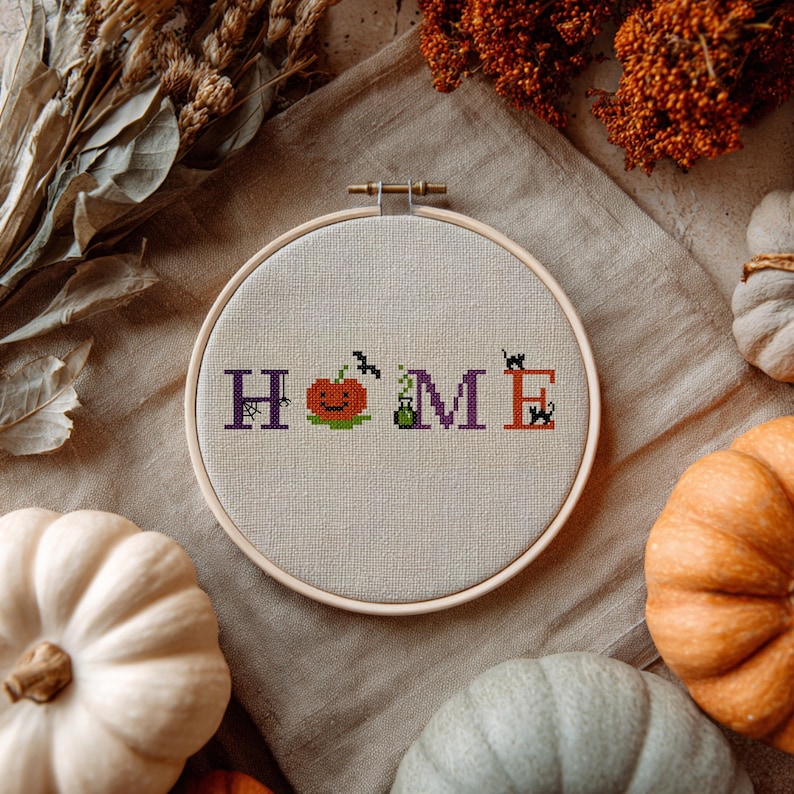 May include: A cross-stitch embroidery hoop with the word "HOME" in purple and orange letters. The "O" is replaced with a smiling jack-o'-lantern. A bat and a black cat are also included in the design. The background is a neutral linen fabric.