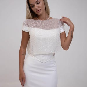 May include: A white bridal gown with a sequined bodice and a fitted skirt. The top has short sleeves and a sheer neckline. The skirt is a sleek, floor-length design. The dress is made of a smooth, satin-like fabric.