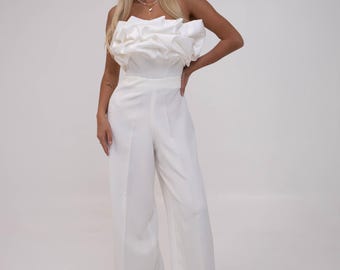 May include: A white strapless jumpsuit with a ruffled bodice and wide-leg pants. The outfit is made of a smooth, flowing fabric. The model is wearing a necklace.