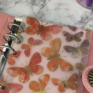 May include: A pink leather bound planner with a clear plastic butterfly patterned insert. The planner has silver rings and a silver metal clasp.