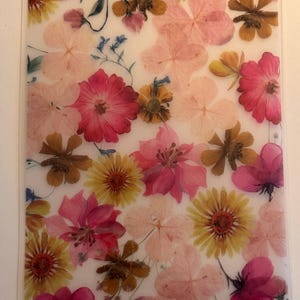 May include: A clear, rectangular piece featuring pressed flowers in shades of pink, yellow, and brown, with small blue accents. The flowers are arranged on a white background. The piece is rectangular.