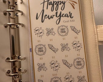 May include: A beige planner with a "Happy New Year" savings tracker. The tracker has a goal of $50 and features illustrations of fireworks, balloons, and rockets. The planner is open, revealing the savings tracker inside.