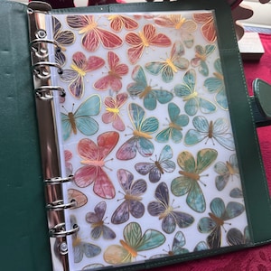 May include: A green leather binder with a clear plastic insert featuring a colorful butterfly pattern. The butterflies are painted in shades of pink, blue, green, and gold.