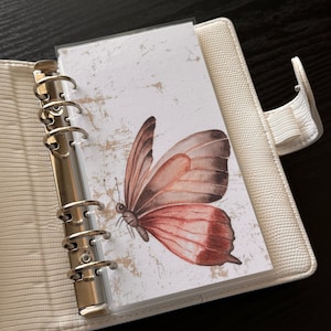 May include: A white binder with a clear plastic pocket containing a watercolor painting of a butterfly with brown and pink wings. The butterfly is on a white background with a distressed texture.