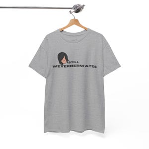 May include: A heather gray t-shirt with a cartoon illustration of a person with black hair and the text "STILL WEVERBERWATES" in black. The shirt is hanging on a wooden hanger.