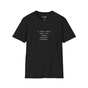 May include: Black t-shirt with the text "I sold feet pics for these concert tickets." in a simple, sans-serif font. The shirt is a basic crew neck style, with short sleeves. The text is centered on the front of the shirt.