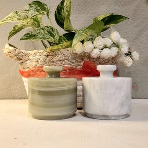 May include: Two cylindrical stone containers with lids, one in a pale sage green with horizontal banding and the other in a white marble finish with grey veining. Both feature a small knob handle on the lid and a smooth, polished surface texture.