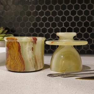 Peut inclure: Un beurrier cylindrique en onyx vert avec un couvercle doté d'une large poignée et une finition polie lisse. La pierre présente des motifs naturels tourbillonnants dans des tons vert clair, crème et brun foncé. Trois petits couteaux en métal.