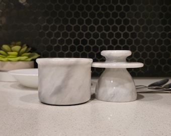 May include: Two white marble butter dishes. The larger dish is a rectangular container with a lid. The smaller dish is a round container with a small, round lid. Both dishes have a marbled pattern.
