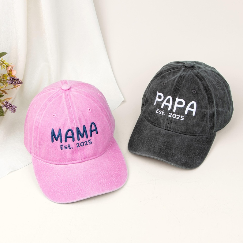 May include: A pink baseball cap with dark blue embroidered text reading MAMA Est. 2025 and a charcoal gray baseball cap with white embroidered text reading PAPA Est. 2025. Both caps feature a structured crown, a curved brim, and eyelet vents.