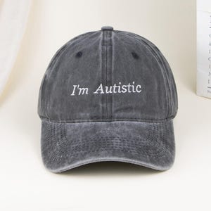 May include: A charcoal grey baseball cap with a washed, distressed finish. The front panel features the white embroidered text "I'm Autistic" in a clean, sans-serif font. The cap has a curved brim and a structured crown with ventilation eyelets.