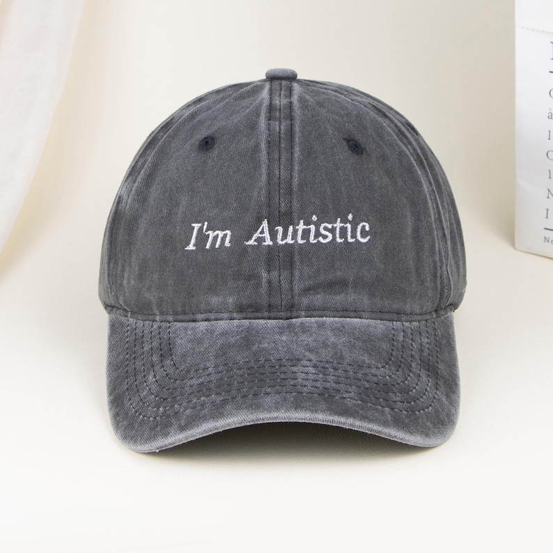 May include: A charcoal grey baseball cap with a washed, distressed finish. The front panel features the white embroidered text "I'm Autistic" in a clean, sans-serif font. The cap has a curved brim and a structured crown with ventilation eyelets.