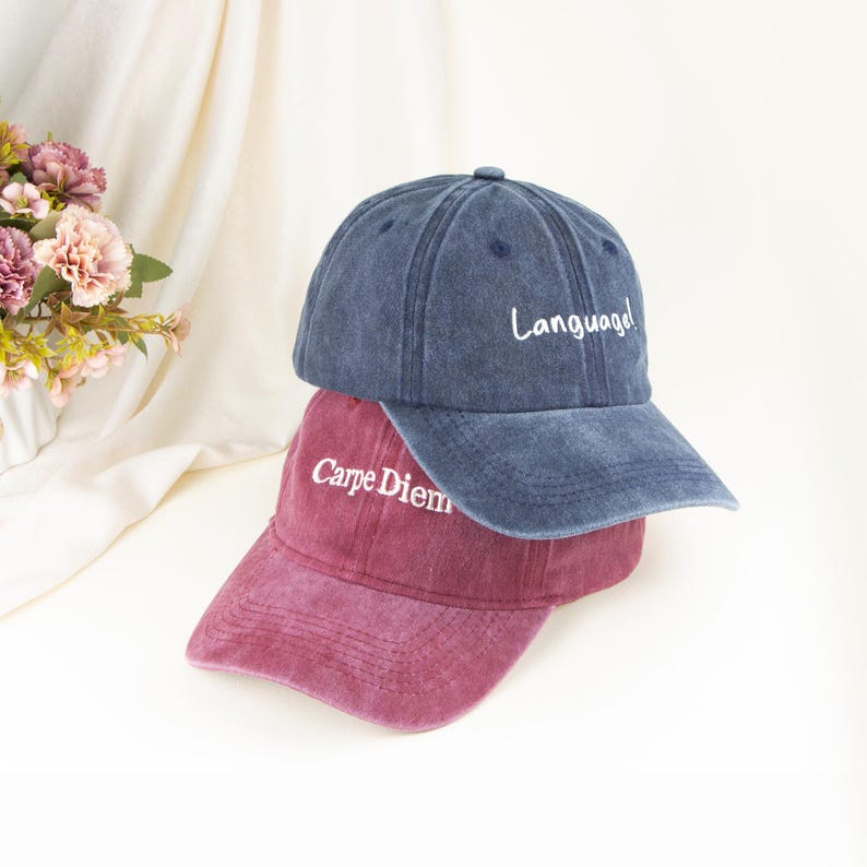 May include: A blue baseball cap with the word Language! embroidered in white and a maroon baseball cap with the word Carpe Diem embroidered in white, both featuring a distressed fabric finish and a curved brim with stitched detailing.
