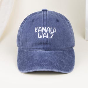 May include: A blue baseball cap with a distressed, washed-out finish. The front of the cap features the white embroidered text KAMALA WALZ in a simple, sans-serif font. The cap has a structured crown, a curved brim, and a button on the top.