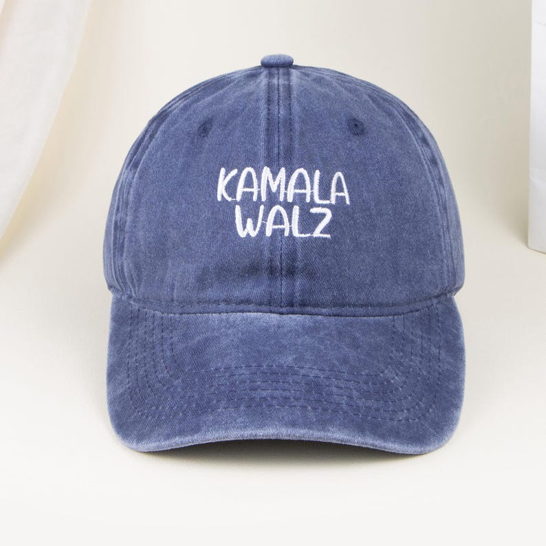 May include: A blue baseball cap with a distressed, washed-out finish. The front of the cap features the white embroidered text KAMALA WALZ in a simple, sans-serif font. The cap has a structured crown, a curved brim, and a button on the top.