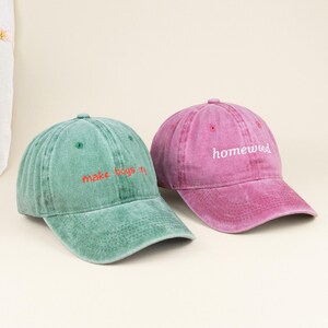 May include: A sage green baseball cap with the red embroidered text make boys cry and a magenta baseball cap with the white embroidered text homewood. Both caps feature a washed, distressed fabric finish, a curved brim, and eyelet vents.