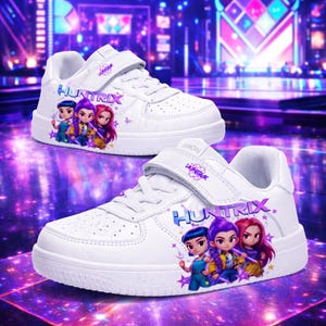 May include: White trainers with a hook-and-loop strap, displaying the word "HUNTRIX" and a cartoon graphic of three characters. The shoes have a perforated design and a purple and blue background.
