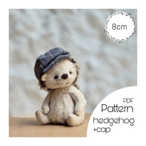Könnte beinhalten: Ein handgefertigtes Igel-Stofftier, etwa 8 cm groß, mit einer blauen Tweed-Mütze. Das Spielzeug hat einen hellbeige Körper, braune Augen und eine dunkelbraune Nase. Der Text "PDF Pattern hedgehog + cap" ist enthalten.