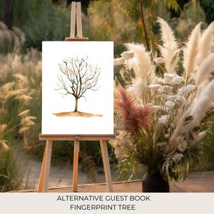 May include: A white canvas with a watercolor painting of a bare tree on an easel. The easel is set up outdoors in front of a bouquet of dried pampas grass. The text "ALTERNATIVE GUEST BOOK FINGERPRINT TREE" is at the bottom of the image.