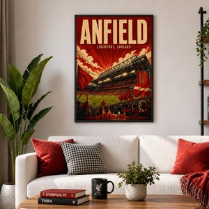 Könnte beinhalten: Ein gerahmter Kunstdruck mit dem Wort "ANFIELD" in cremefarbenen Buchstaben über "LIVERPOOL, ENGLAND". Das Kunstwerk zeigt eine Stadionszene in Rot- und Orangetönen mit einer Menschenmenge und Flaggen. Der Druck hängt über einem weißen Sofa mit roten Kissen.