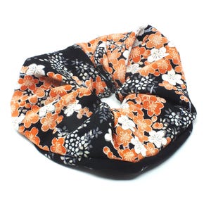 May include: A black scrunchie with a floral pattern in orange and white. The fabric is folded and gathered, creating a textured appearance. The scrunchie is designed to hold hair.