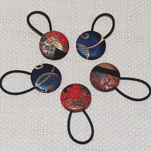 May include: Five round hair ties with decorative fabric covers. The fabric features patterns in red, gold, and blue. Each tie has a black elastic band. The ties are arranged in a circular pattern.