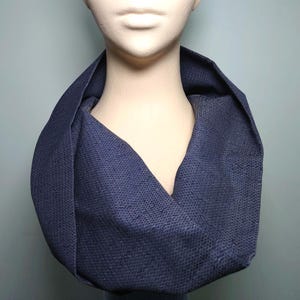 以下が含まれることがあります： ネイビーブルーのテクスチャード加工生地で作られたインフィニティスカーフ。マネキンの首に巻き付けられ、デザインを示しています。生地には微妙な織り模様があり、アクセサリーに視覚的な魅力を加えています。