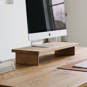 May include: A rectangular wooden monitor stand with a natural grain finish, designed to elevate a computer screen. The stand features a flat top surface supported by two solid side panels, providing a stable base for a desktop computer monitor.
