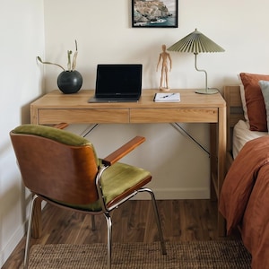 Könnte beinhalten: Ein Schreibtisch aus Holz mit einem Laptop, einem kleinen gerahmten Landschaftsfoto, einer Holzpuppe und einer grünen Plisseelampe. Ein Stuhl mit brauner Lederrückenlehne und grünem Sitz steht vor dem Schreibtisch neben einem Bett mit rostfarbener und hellblauer Bettwäsche.