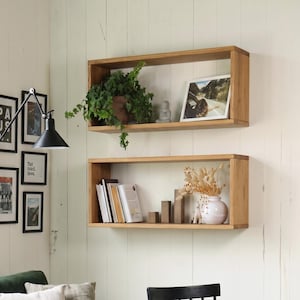 May include: Two rectangular light wood wall shelves holding a green plant, a framed photograph, books, wooden blocks, and a white vase with dried flowers. The shelves are mounted on a white panelled wall above a dark green velvet sofa.