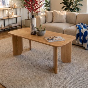 May include: A rectangular light wood coffee table with curved legs, featuring a tall textured vase with red berries, a small gray bowl with cacti, and a magazine on top. The table sits on a light gray textured area rug in a bright living room.