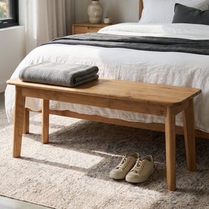 May include: A rectangular wooden bench with four tapered legs, featuring a light brown wood grain finish. A folded grey textured blanket rests on the seat of the bench, which is positioned at the foot of a bed with white linens and pillows.