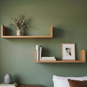 May include: Two light-toned wooden U-shaped wall shelves mounted on a sage green wall. The top shelf holds a small vase with dried foliage. The bottom shelf displays three books and a framed abstract print. A wooden dresser and bed are below.