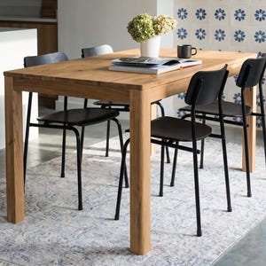 Puede incluir: Una mesa de comedor rectangular de madera clara con cuatro sillas negras de patas metálicas finas y respaldos curvos. Sobre la mesa hay un libro abierto, un jarrón blanco con flores verdes y una taza negra, todo sobre una alfombra estampada.