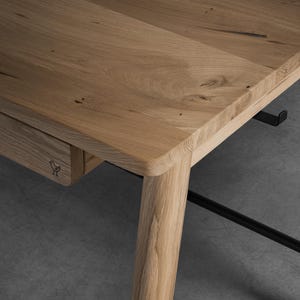 May include: A light brown oak wood desk featuring a smooth rectangular top, rounded corners, and a single drawer. The drawer front is marked with a small black bird icon. The desk is supported by sturdy wooden legs and includes a black metal frame.