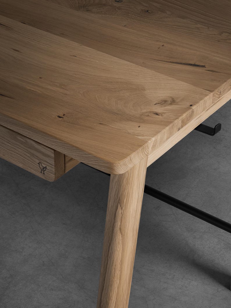 May include: A light brown oak wood desk featuring a smooth rectangular top, rounded corners, and a single drawer. The drawer front is marked with a small black bird icon. The desk is supported by sturdy wooden legs and includes a black metal frame.