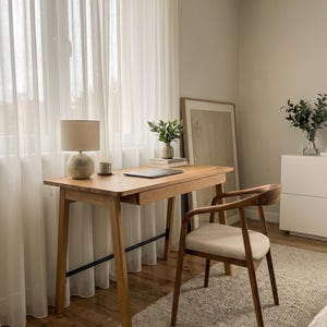 May include: A light wood desk with a single drawer and a black metal support bar, paired with a matching wood chair featuring a beige upholstered seat. On the desk sits a round lamp with a beige shade, a small cup, and a closed laptop.