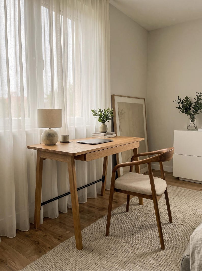 May include: A light wood desk with a single drawer and a black metal support bar, paired with a matching wood chair featuring a beige upholstered seat. On the desk sits a round lamp with a beige shade, a small cup, and a closed laptop.